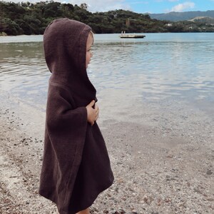 Puede incluir: Un poncho con capucha de color marr&oacute;n oscuro que presenta un tejido de punto de gofre texturizado. La prenda tiene un dise&ntilde;o holgado y sin mangas con una capucha que cubre la cabeza y cae hasta la mitad del muslo, ofreciendo una capa simple.