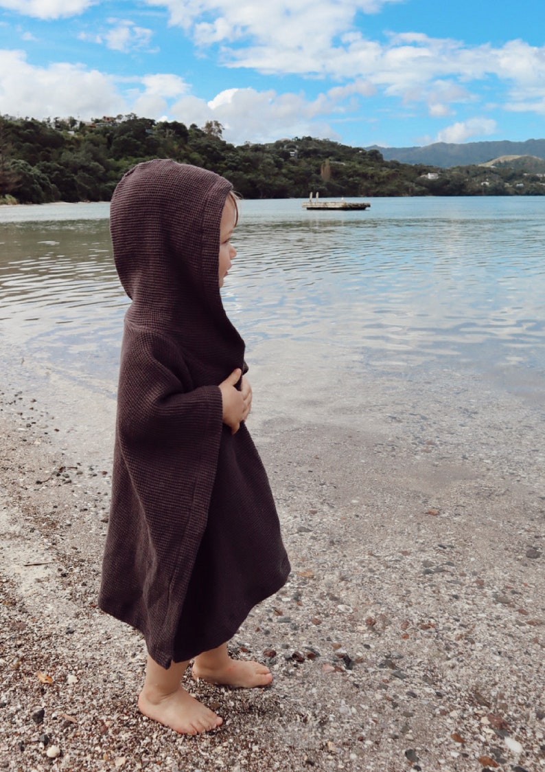 Puede incluir: Un poncho con capucha de color marr&oacute;n oscuro que presenta un tejido de punto de gofre texturizado. La prenda tiene un dise&ntilde;o holgado y sin mangas con una capucha que cubre la cabeza y cae hasta la mitad del muslo, ofreciendo una capa simple.
