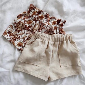 May include: Two pairs of shorts. One pair is beige linen with pockets and an elastic waistband. The other pair is brown and white floral with a ruffled waistband.