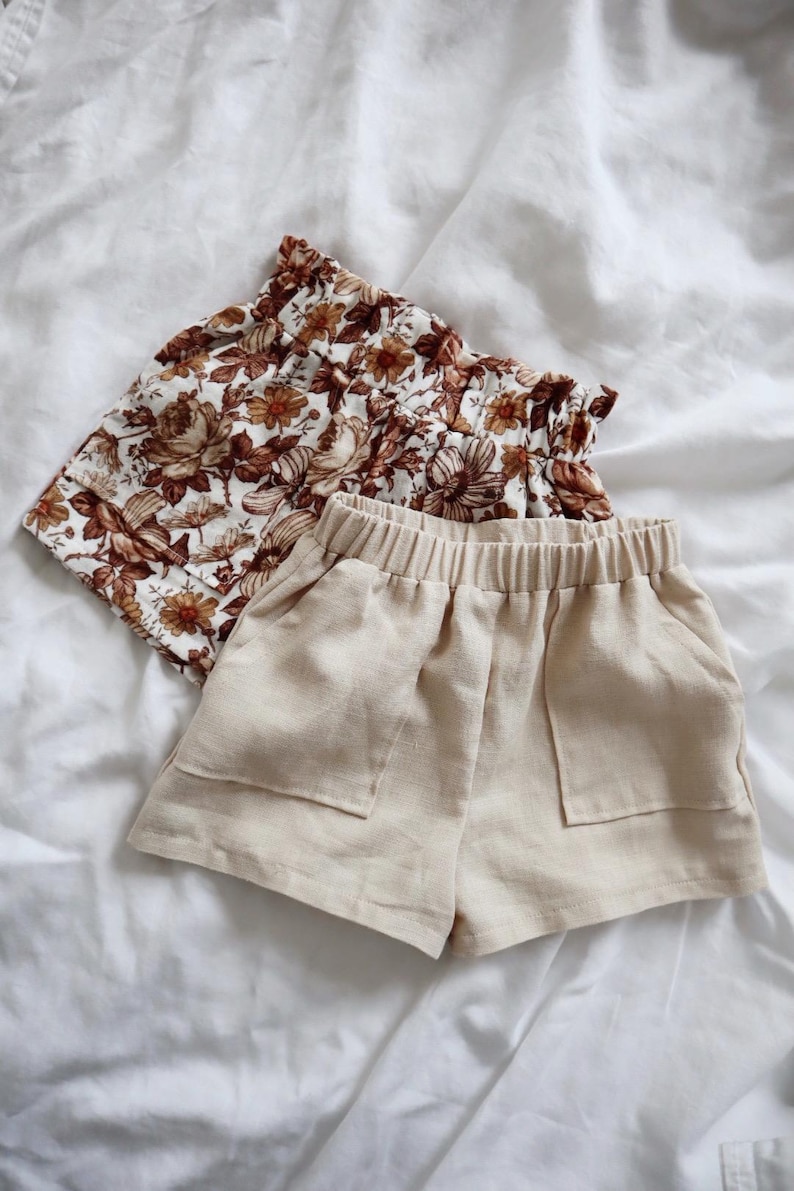 May include: Two pairs of shorts. One pair is beige linen with pockets and an elastic waistband. The other pair is brown and white floral with a ruffled waistband.