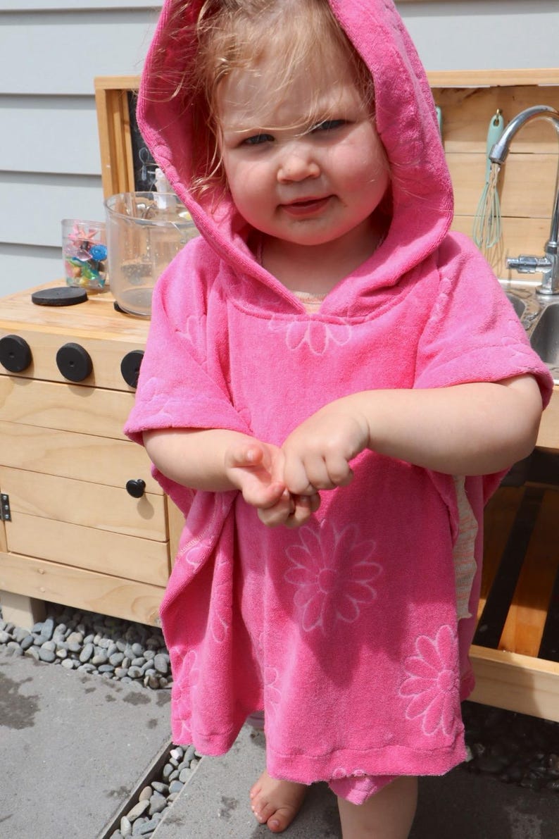 Puede incluir: Un poncho de toalla con capucha de color rosa brillante que presenta un patr&oacute;n repetitivo de margaritas blancas. La prenda est&aacute; confeccionada en un material de felpa suave y absorbente, con capucha, mangas cortas y un corte holgado.