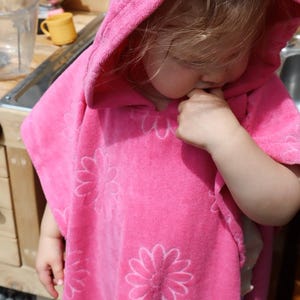 Puede incluir: Un poncho de toalla con capucha en color rosa brillante que presenta un patr&oacute;n floral en relieve por toda la prenda. El tejido suave y absorbente incluye una capucha amplia y aberturas anchas para los brazos, brindando calidez y confort.