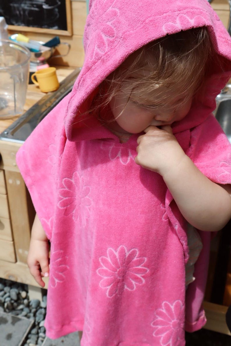 Puede incluir: Un poncho de toalla con capucha en color rosa brillante que presenta un patr&oacute;n floral en relieve por toda la prenda. El tejido suave y absorbente incluye una capucha amplia y aberturas anchas para los brazos, brindando calidez y confort.