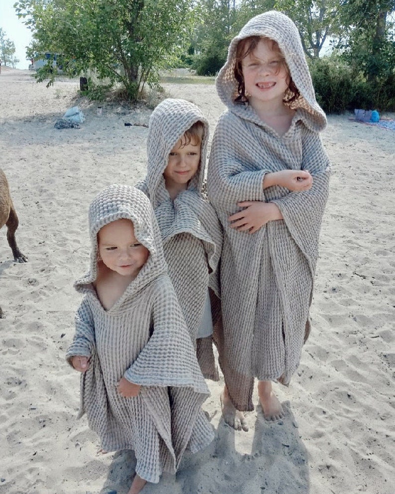 Puede incluir: Tres ponchos con capucha confeccionados en tela de color beige claro con textura de gofre. Cada poncho presenta un corte holgado y drapeado con mangas anchas y una capucha profunda. El material texturizado tiene un patr&oacute;n de cuadr&iacute;cula.