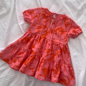 May include: A pink and orange floral print dress with a ruffled skirt and short sleeves. The dress has a tie at the back of the neck.