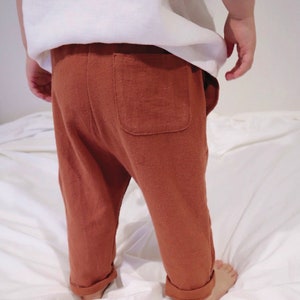 以下が含まれることがあります： 錆色の幼児用パンツ。背面にポケットが1つ、裾は折り返されています。白いトップスと組み合わせています。子供は白い表面に立っています。