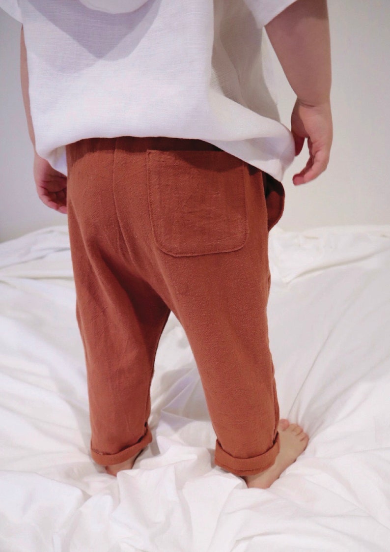 以下が含まれることがあります： 錆色の幼児用パンツ。背面にポケットが1つ、裾は折り返されています。白いトップスと組み合わせています。子供は白い表面に立っています。
