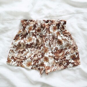 May include: White linen shorts with a brown floral print. The shorts have an elastic waistband and a ruffled hem.