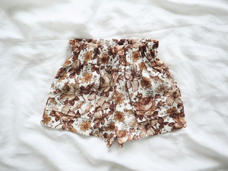 May include: White linen shorts with a brown floral print. The shorts have an elastic waistband and a ruffled hem.