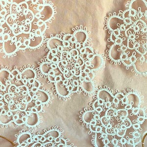 May include: Close-up of several white lace doilies with intricate floral patterns. The doilies are arranged on a light pink surface, with gold-colored threads visible. The delicate lace features a detailed, openwork design, perfect for crafting or home decor.