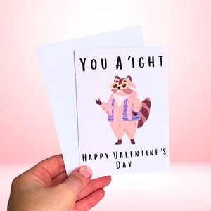 May include: A Valentine's Day card with a cartoon raccoon wearing a pink jacket and sunglasses. The card reads "YOU A'IGHT HAPPY VALENTINE'S DAY" in black text. A white envelope is behind the card.