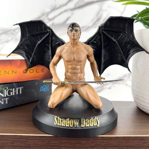 Puede incluir: Una figura de un hombre arrodillado con grandes alas negras de murci&eacute;lago, cabello oscuro y maquillaje azul en los ojos. La figura sostiene una espada plateada delgada horizontalmente sobre el regazo. La base circular negra tiene el texto Shadow Daddy.