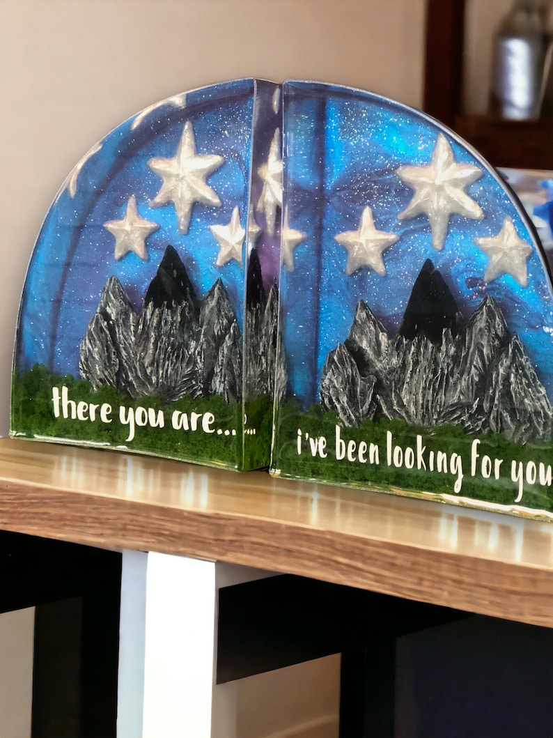 May include: A pair of bookends with a blue and purple galaxy design with white stars and black mountains. The left bookend says "there you are..." and the right bookend says "i've been looking for you".
