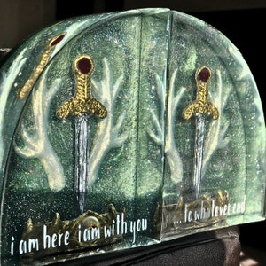 May include: A pair of bookends with a green glitter background and white antlers. Each bookend features a gold sword with a red jewel and the text "i am here i am with you... to whatever end".