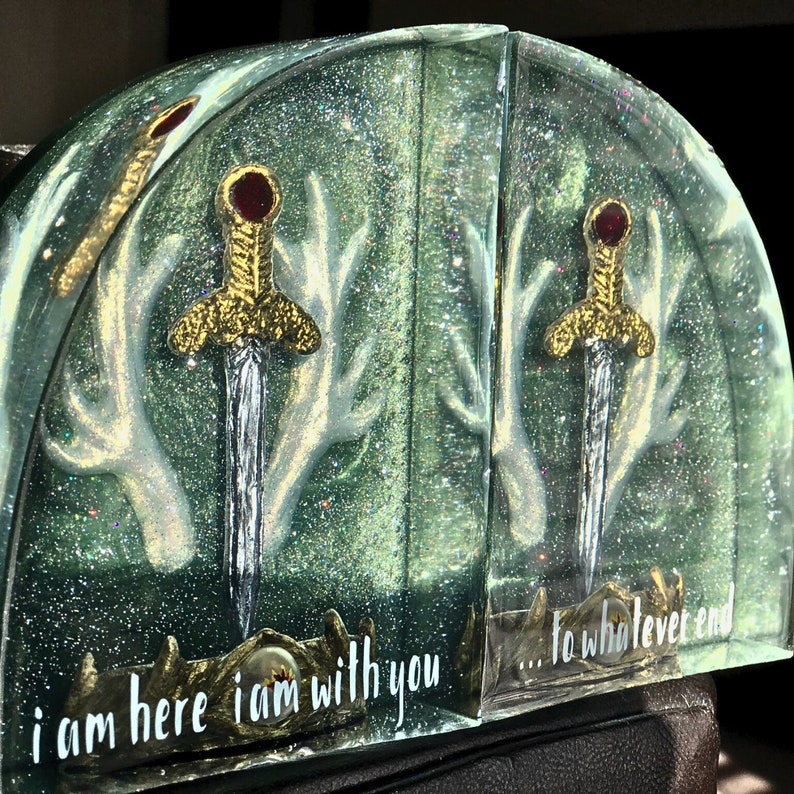 May include: A pair of bookends with a green glitter background and white antlers. Each bookend features a gold sword with a red jewel and the text "i am here i am with you... to whatever end".