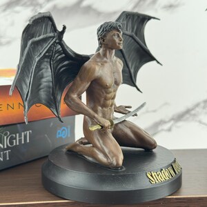 Puede incluir: Una figura de tono bronce de un personaje alado arrodillado con cabello oscuro y musculatura definida, sosteniendo una peque&ntilde;a espada. La figura tiene grandes alas negras texturizadas y est&aacute; sobre una base circular negra con el texto dorado Shadow Hunt.