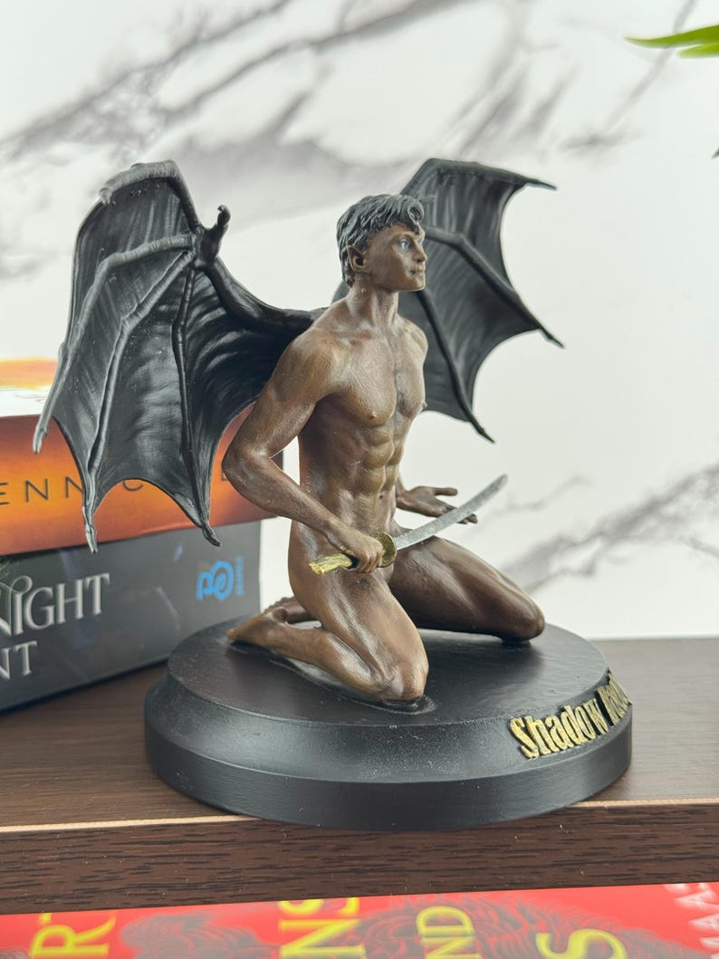 Puede incluir: Una figura de tono bronce de un personaje alado arrodillado con cabello oscuro y musculatura definida, sosteniendo una peque&ntilde;a espada. La figura tiene grandes alas negras texturizadas y est&aacute; sobre una base circular negra con el texto dorado Shadow Hunt.