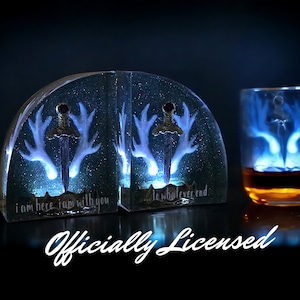May include: A pair of bookends with a glowing blue design of antlers and a sword. The bookends are made of clear resin and have the text "I am here I am with you... to whatever end" on them.