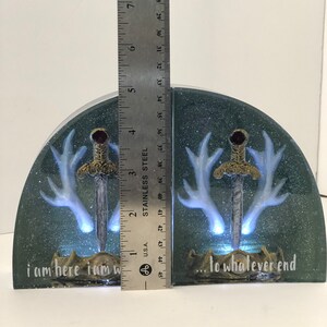 May include: A pair of bookends with a dark green background and glitter. Each bookend features a white deer antler with a silver sword in the center. The text "i am here i am w... to whatever end" is written on the bottom of the bookends.