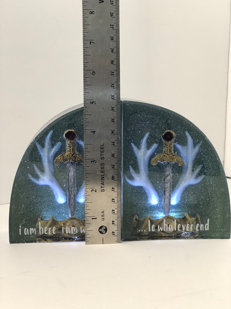 May include: A pair of bookends with a dark green background and glitter. Each bookend features a white deer antler with a silver sword in the center. The text "i am here i am w... to whatever end" is written on the bottom of the bookends.