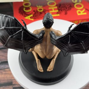 Puede incluir: Una figura con cabello oscuro, piel bronceada y grandes alas negras similares a las de un murci&eacute;lago, arrodillada sobre una base circular negra. La figura se muestra desde atr&aacute;s, revelando la estructura detallada de las alas.