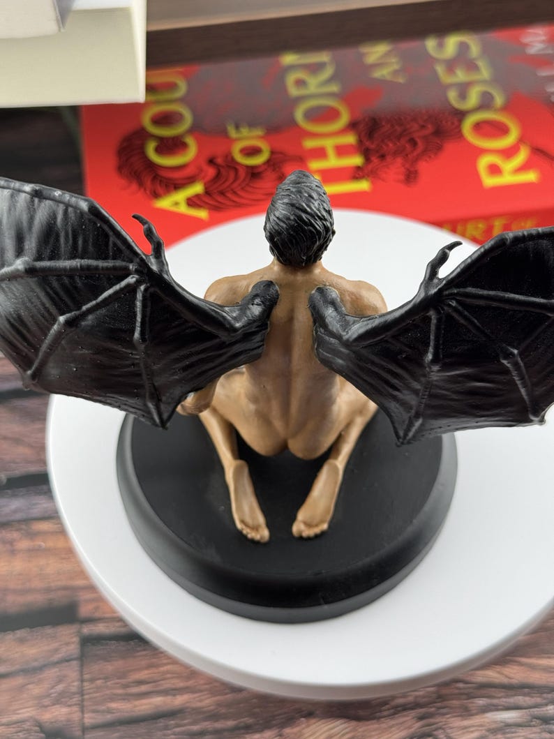Puede incluir: Una figura con cabello oscuro, piel bronceada y grandes alas negras similares a las de un murci&eacute;lago, arrodillada sobre una base circular negra. La figura se muestra desde atr&aacute;s, revelando la estructura detallada de las alas.