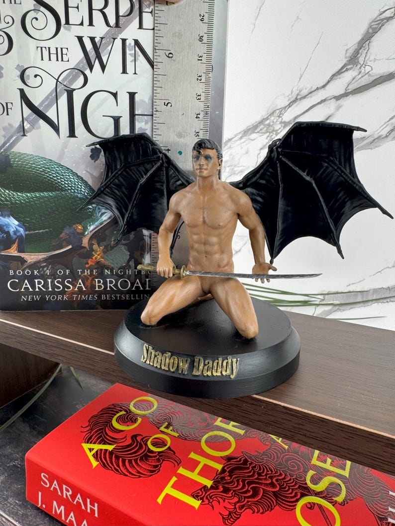 Puede incluir: Una figura masculina de piel oscura, alas negras y una espada, arrodillada sobre una base circular negra con la inscripci&oacute;n Shadow Daddy. Una regla met&aacute;lica detr&aacute;s de la figura muestra una altura de unos 15 cm. Situada frente a un libro.