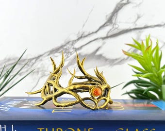 Puede incluir: Una corona de color dorado con un diseño texturizado similar a ramas o astas entrelazadas. La corona está acentuada con una piedra preciosa circular de tono naranja engastada en la parte frontal, creando un punto focal en medio de la estructura metálica de aspecto orgánico.