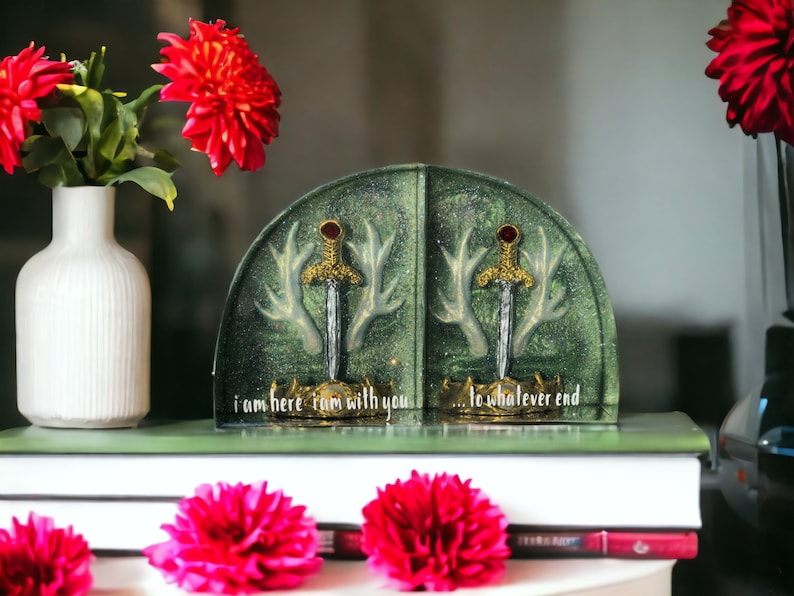 May include: A pair of bookends featuring a green and gold design with swords and antlers. The bookends are sitting on a stack of books. The text on the bookends reads "I am here I am with you... to whatever end."