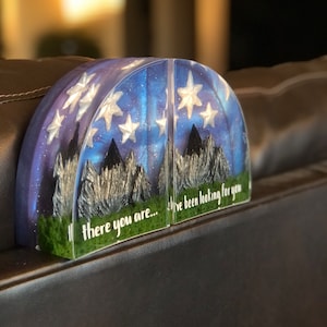 May include: A pair of bookends with a galaxy and mountain scene. The bookends are made of clear resin with glitter and silver stars. The text on the bookends reads "there you are... I've been looking for you".
