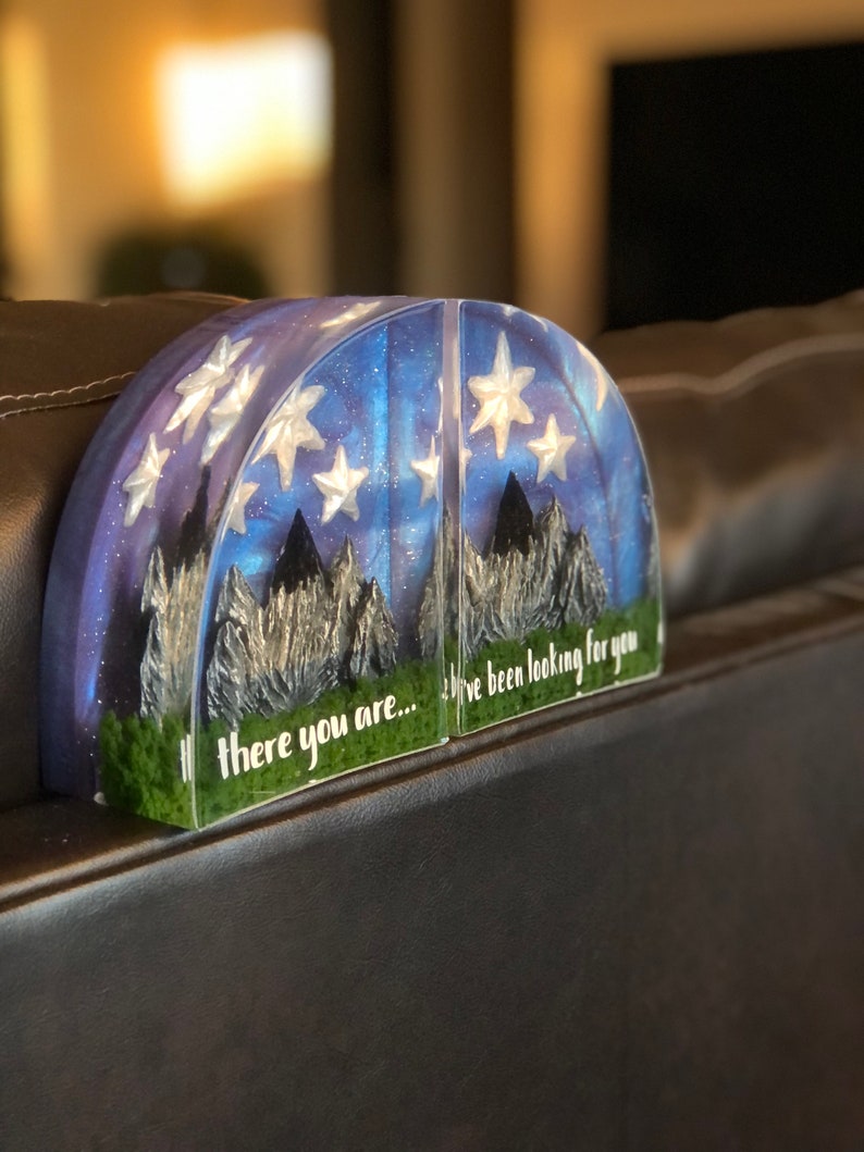 May include: A pair of bookends with a galaxy and mountain scene. The bookends are made of clear resin with glitter and silver stars. The text on the bookends reads "there you are... I've been looking for you".