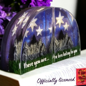 May include: A pair of bookends shaped like a crescent moon with a starry night sky and mountain silhouette. The bookends are made of clear resin and have a glittery finish. The text "there you are... I've been looking for you" is written on the bookends.