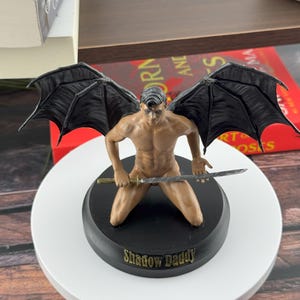 Puede incluir: Una figura de un personaje arrodillado con grandes alas negras, sosteniendo una espada horizontalmente. La figura tiene un tono de piel bronceado y cabello oscuro. Est&aacute; montada sobre una base negra redonda con el texto Shadow Daddy en letras doradas.
