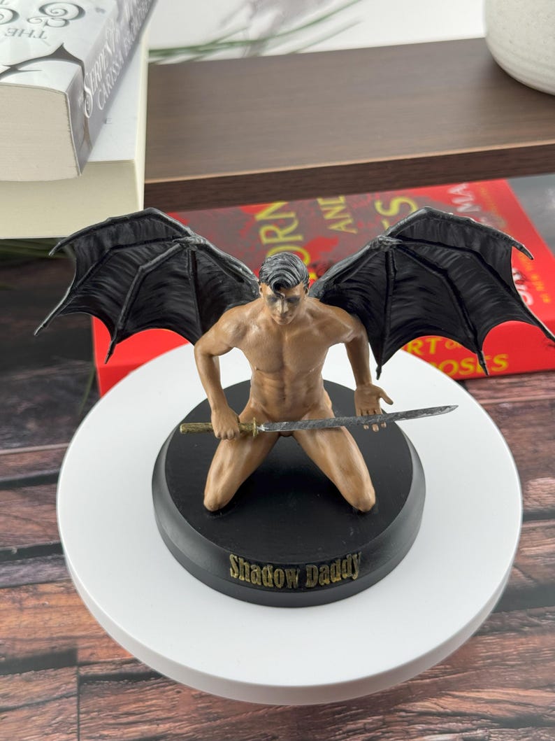 Puede incluir: Una figura de un personaje arrodillado con grandes alas negras, sosteniendo una espada horizontalmente. La figura tiene un tono de piel bronceado y cabello oscuro. Est&aacute; montada sobre una base negra redonda con el texto Shadow Daddy en letras doradas.