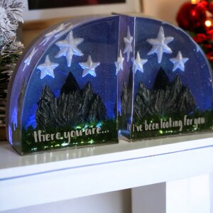 May include: A pair of bookends made of clear resin with a blue and white starry night sky design. The bookends feature a silhouette of mountains and the text "there you are..." and "I've been looking for you".