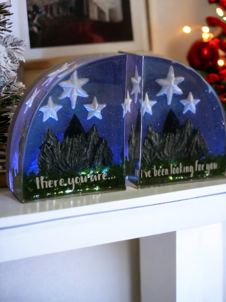 May include: A pair of bookends made of clear resin with a blue and white starry night sky design. The bookends feature a silhouette of mountains and the text "there you are..." and "I've been looking for you".