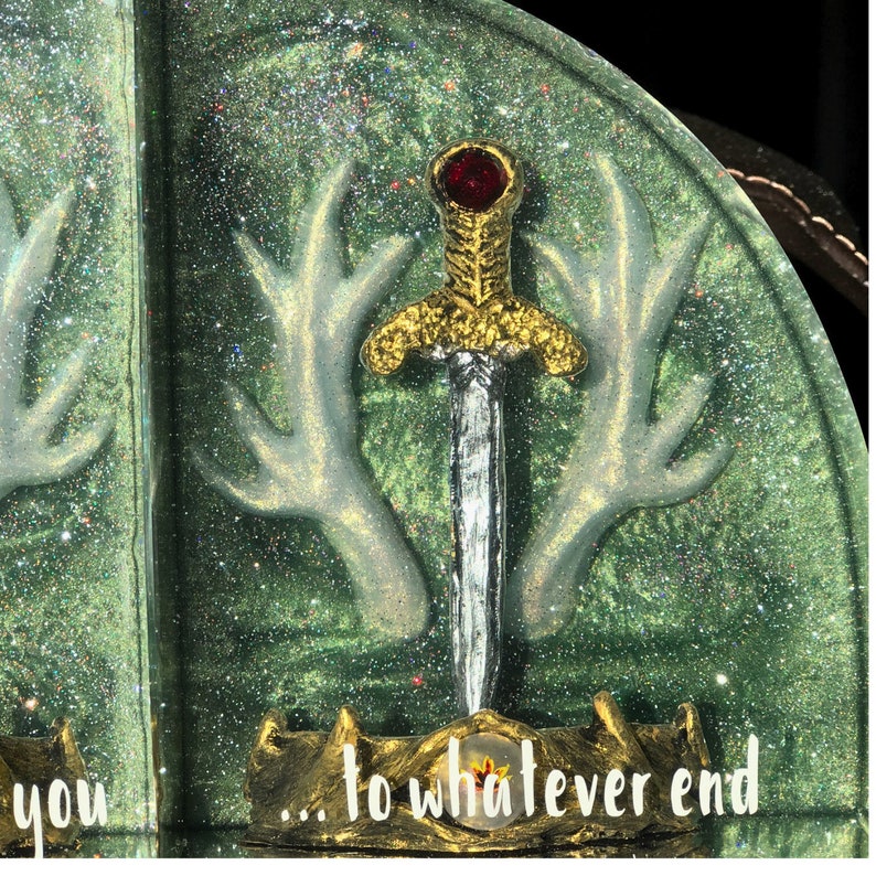May include: A green and white bookend with a gold sword and red jewel. The bookend features a pair of antlers and the text "...to whatever end".