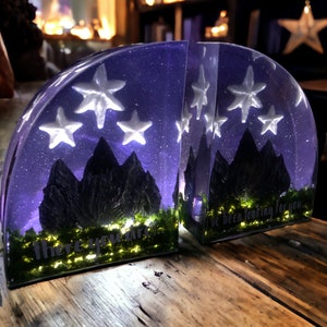 May include: Two bookends made of clear resin with a purple and black mountain scene and white stars. The bookends are lit from within and the text "there you are..." and "I've been looking for you" are visible on the front of each bookend.