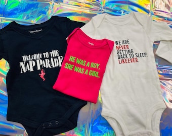 May include: Three baby onesies with text. A navy blue onesie says "WELCOME TO THE NAP PARADE". A pink onesie says "HE WAS A BOY, SHE WAS A GIRL". A white onesie says "WE ARE NEVER GETTING BACK TO SLEEP. LIKEEVER."