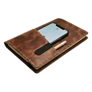 Puede incluir: Un cuaderno de cuero marrón con un teléfono inteligente en el bolsillo y un lápiz. El cuaderno tiene una superficie texturizada y una banda elástica negra para sujetar el teléfono. El lápiz es de madera con una goma de borrar rosa.