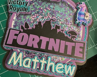 Könnte beinhalten: Lila und schwarzer Fortnite-Kuchentopper mit glitzernder Oberfläche. Der Topper zeigt die Wörter "Fortnite" und "Matthew" sowie ein Lama und die Nummer 12. Der Topper enthält auch die Wörter "#1 Victory Royale".