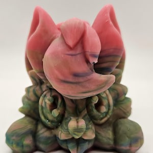 Könnte beinhalten: Eine kleine, handbemalte Keramikskulptur einer Kreatur mit rosa Ohren, einem grün-rosa Körper und einem Wirbelmuster.