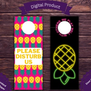 Puede incluir: Colgadores de puerta con temática de piña. Uno presenta un fondo rosa con piñas amarillas repetidas y el texto "PLEASE DISTURB US". El otro tiene un fondo negro con un diseño de piña de neón. Dimensiones: 11 x 28 cm.