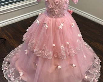 May include: A pink off-the-shoulder dress with a layered tulle skirt, decorated with floral appliqués and a lace trim. A small crown sits atop the dress form. The dress is designed for a special occasion.