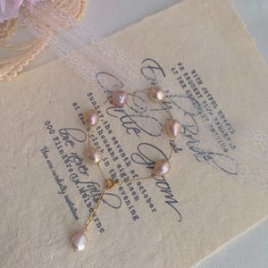 May include: A delicate pearl bracelet with a gold chain and a single pearl charm hanging from the end. The bracelet is on a cream-coloured background with a lace overlay and text that reads "Sunday, the seventh of October at six o'clock in the evening" and "Love never fails".