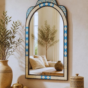 May include: An arched wall mirror with a decorative blue and white geometric pattern. The mirror has a dark frame and is hanging on a white wall. The mirror reflects a room with a couch, a wooden table, and a vase.