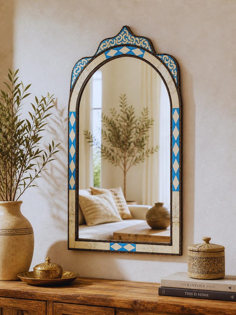 May include: An arched wall mirror with a decorative blue and white geometric pattern. The mirror has a dark frame and is hanging on a white wall. The mirror reflects a room with a couch, a wooden table, and a vase.