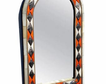 May include: An arched mirror with a decorative frame. The frame features a pattern of orange, silver, and white diamond shapes, bordered by a cream-colored trim. The mirror has a brass-colored edge.