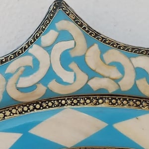 May include: Close-up of a decorative plate with a turquoise and white geometric design. The plate features a star-shaped edge with a detailed border and inlaid white shapes. The design is symmetrical and ornate.