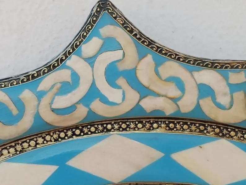 May include: Close-up of a decorative plate with a turquoise and white geometric design. The plate features a star-shaped edge with a detailed border and inlaid white shapes. The design is symmetrical and ornate.